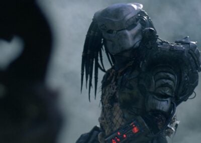 A imagem mostra um Predador, com a sua icónica armadura de batalha e máscara, do filme O Predador (lançado em 2018).